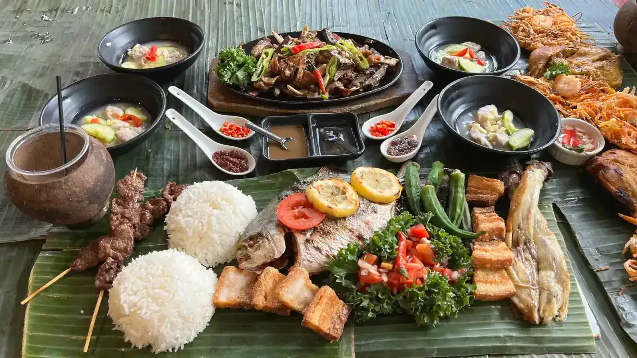 Kamayan feast spread — sisig, BBQ skewers, fried fish, rice mounds, soup, and coconut drinks on banana leaves